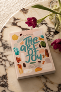 A book titled "The Art of Joy" by Jen Sievers lies on a white marble tray, styled with two purple tulips in a glass vase on a textured cream fabric background.