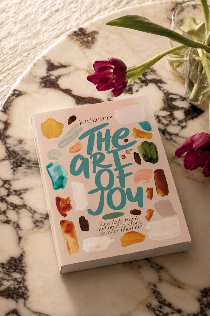 A book titled "The Art of Joy" by Jen Sievers lies on a white marble tray, styled with two purple tulips in a glass vase on a textured cream fabric background.