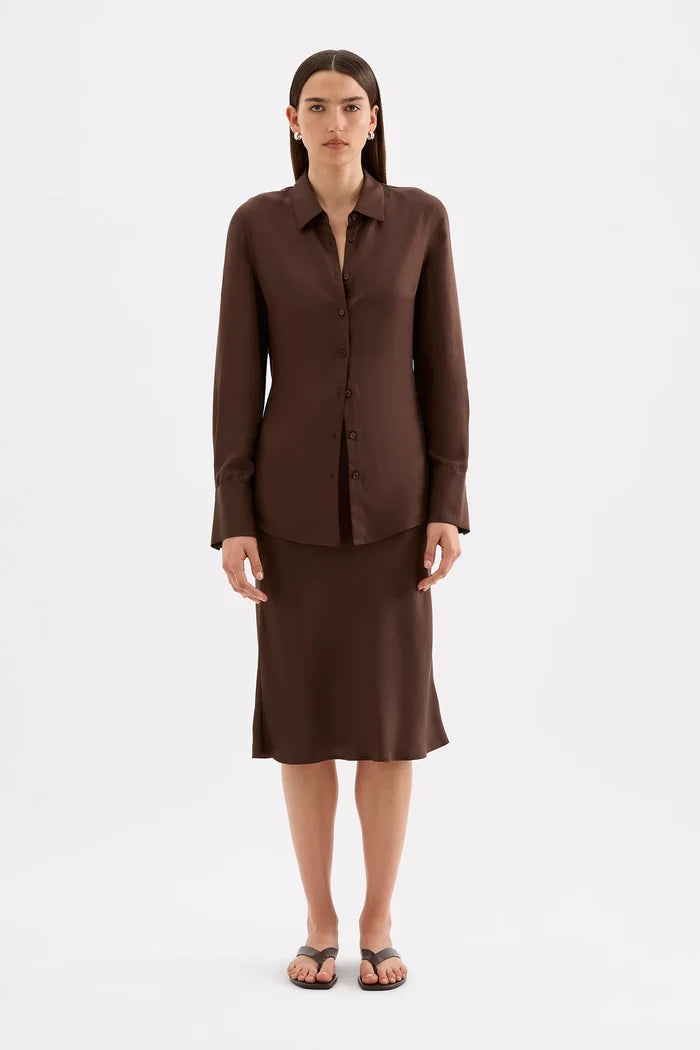 A woman stands against a plain light gray studio background wearing a matching dark brown button-front blouse and knee-length skirt set with black flip-flop sandals.