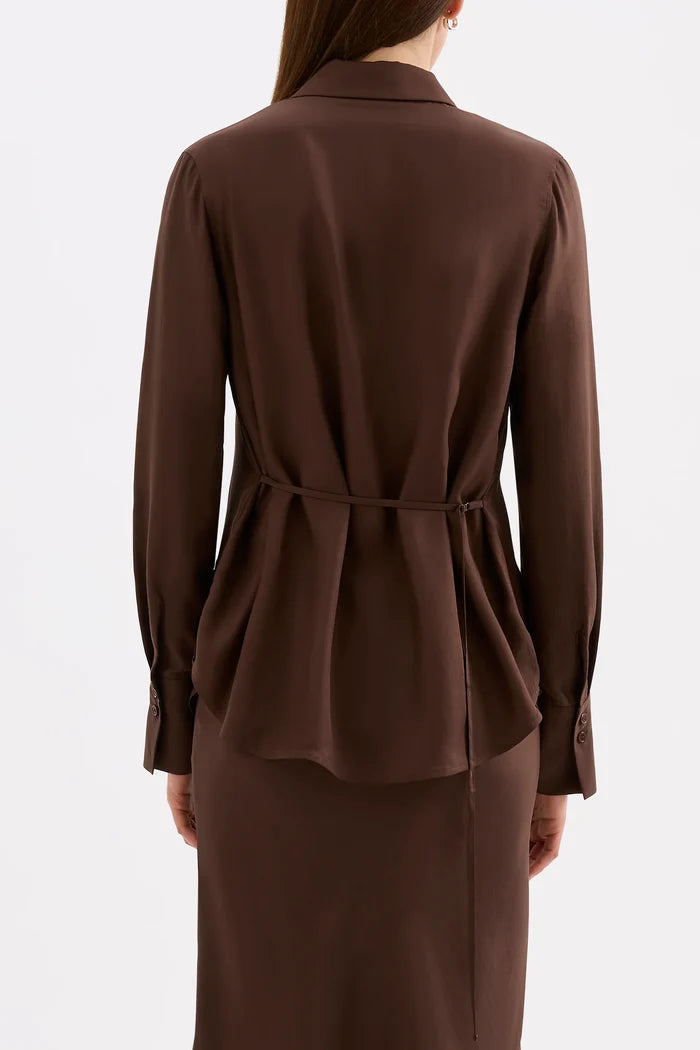 A model shown from the back wearing a dark brown long-sleeve collared blouse with a tie waist and a matching dark brown skirt, set against a plain light gray studio background.