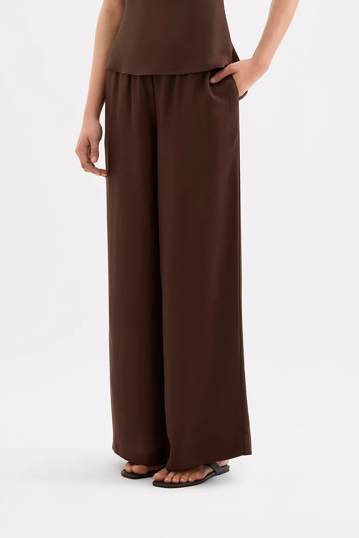 A model wearing dark brown wide-leg trousers with side pockets, paired with a matching dark brown top and black sandals, shown from the waist down against a light gray studio background.