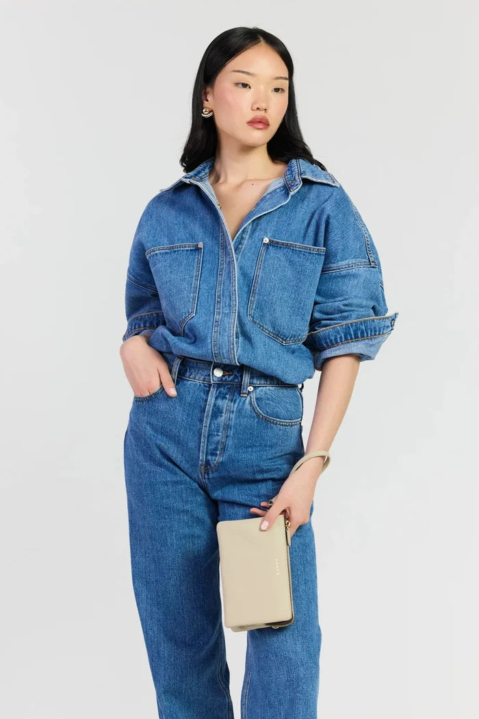 A model wearing a blue denim shirt-jacket and matching blue denim jeans, holding a beige clutch bag against a light gray studio background.
