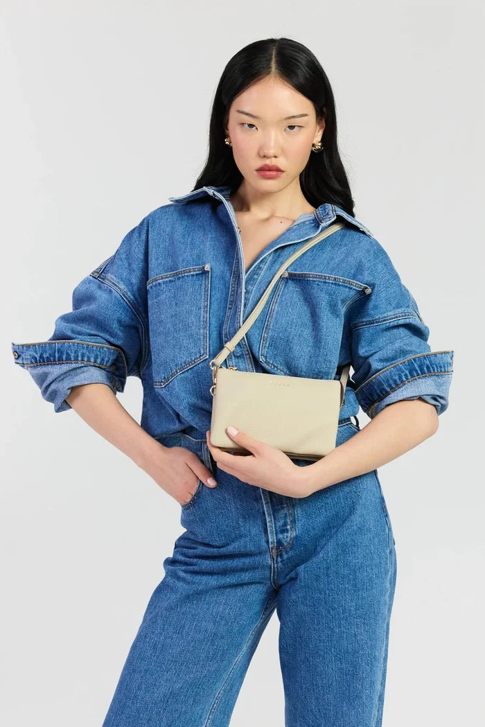 A model wearing a blue denim jumpsuit and matching denim jacket, holding a beige leather crossbody bag with a matching strap against a light gray studio background.