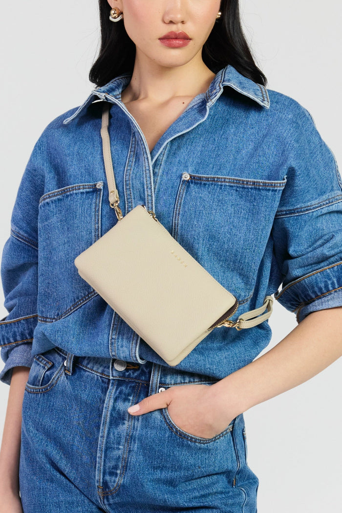 A woman wearing a blue denim shirt and matching denim pants carries a beige leather crossbody bag with a matching strap and gold-tone hardware against a light gray studio background.