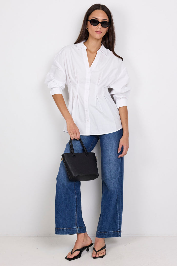 A woman modeling a white oversized button-up shirt with blue wide-leg jeans, black sandals, and a black handbag against a plain white studio background.