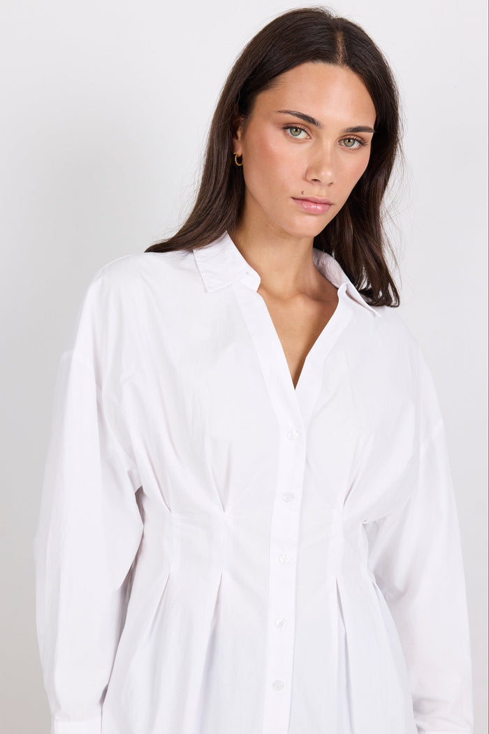 A woman wearing a white long-sleeve button-up shirt dress with a tie waist, photographed against a light gray studio background.