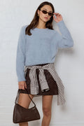 A woman modeling a light blue fuzzy long-sleeve sweater, a brown mini skirt with a black and white striped shirt tied around the waist, and a dark brown leather shoulder bag, standing against a white brick wall.