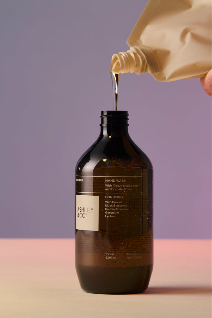 Bottle of Ashley & Co. product with liquid being poured from a beige container against a gradient background.