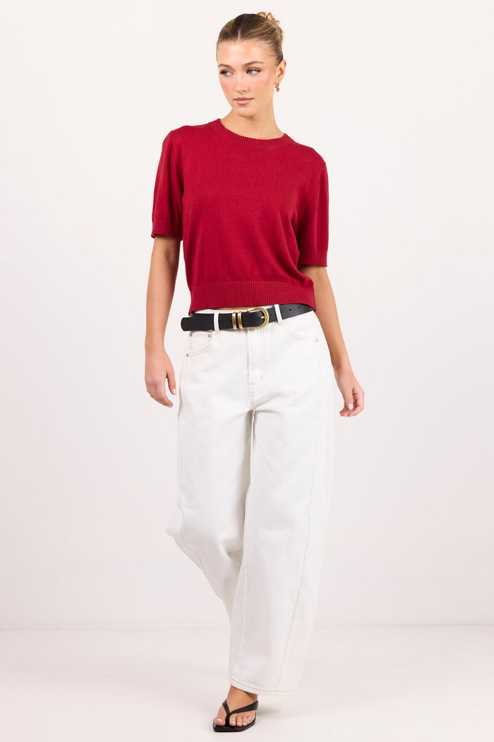 A woman modeling a short-sleeve crewneck knit top in deep red, paired with white wide-leg pants, a black belt, and black sandals, against a plain white studio background.