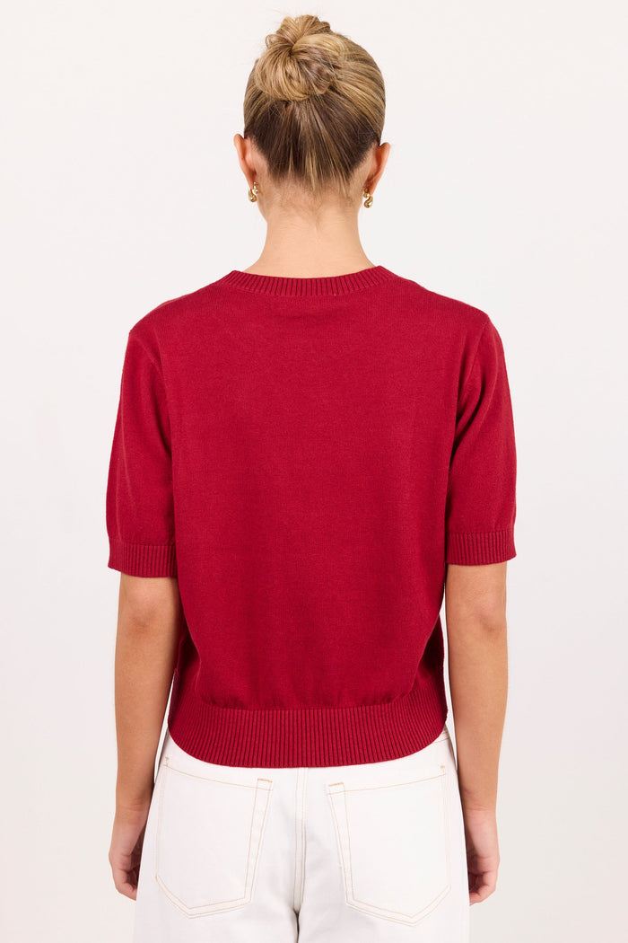 A woman viewed from behind wearing a short-sleeve knit sweater in deep red and white pants, standing against a plain white studio background.