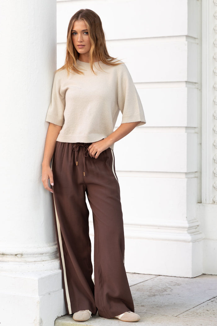 Woman wearing a beige top and brown pants standing against a white wall.