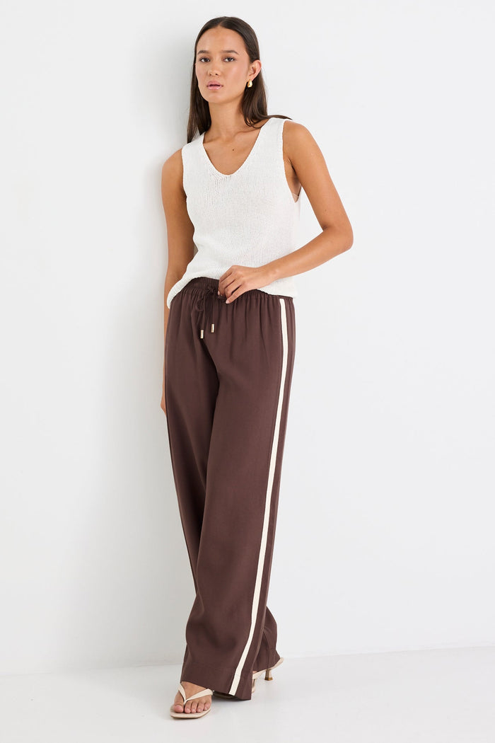 Woman wearing a white sleeveless top and brown pants with a white stripe on a white background