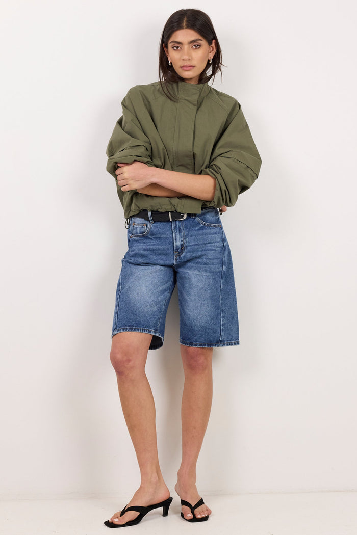 A model stands against a plain white studio background wearing an olive green cropped jacket, blue denim shorts, and black heeled sandals.