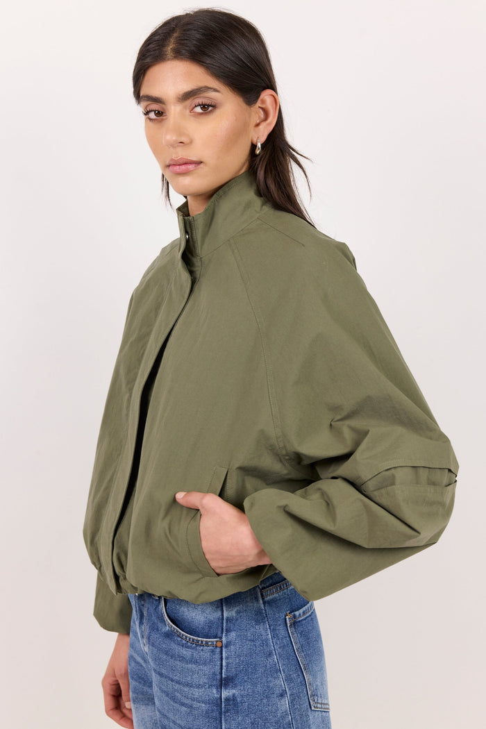 A woman wearing an olive green cropped utility jacket with a high collar and voluminous sleeves, paired with blue jeans, photographed against a plain off-white studio background.