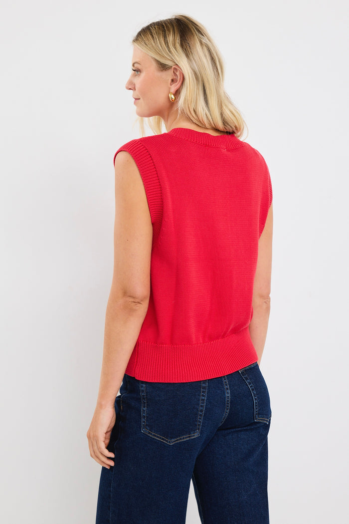 Woman wearing a sleeveless red knit vest with gold buttons down the front, styled with dark denim and loose blonde waves.