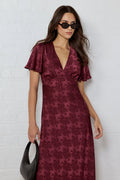 Woman wearing a burgundy patterned dress against a white brick wall.