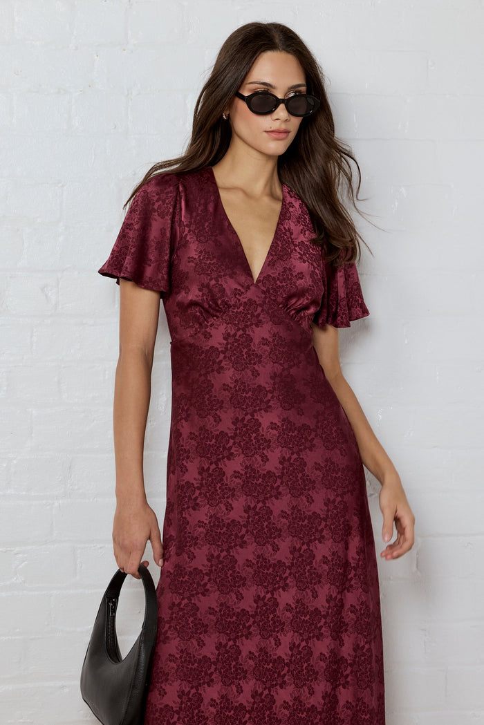 Woman wearing a burgundy patterned dress against a white brick wall.