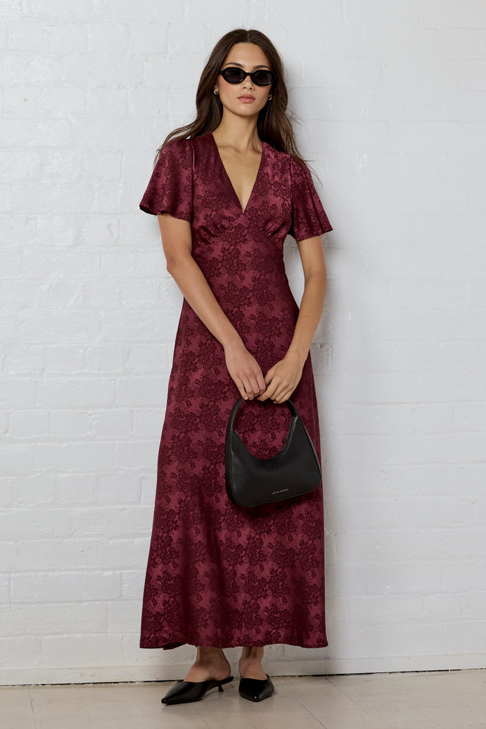 Woman wearing a burgundy patterned dress holding a black handbag against a white brick wall.