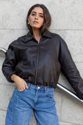 Woman wearing a black leather jacket and blue jeans against a gray wall.