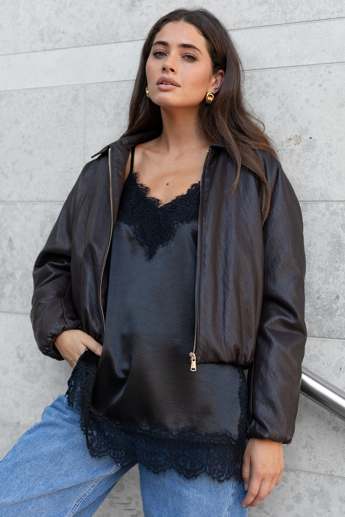 Woman wearing a black lace top and leather jacket against a gray wall.