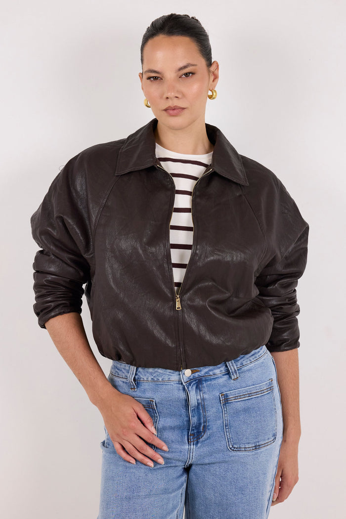 A model wearing a dark brown leather jacket with a zippered front, styled over a black and white striped t-shirt and light blue jeans, against a plain light gray background.