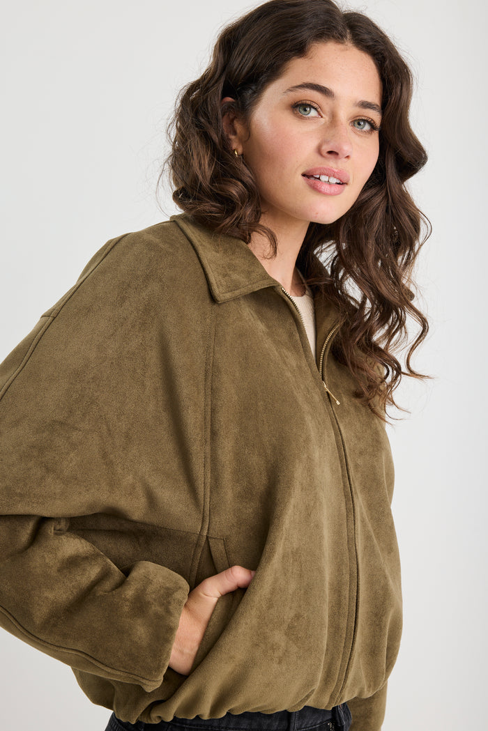 Woman wearing a khaki mock suede zip-up jacket with a soft, textured finish and a relaxed fit, styled over a beige knit top and black jeans.
col_pattern
