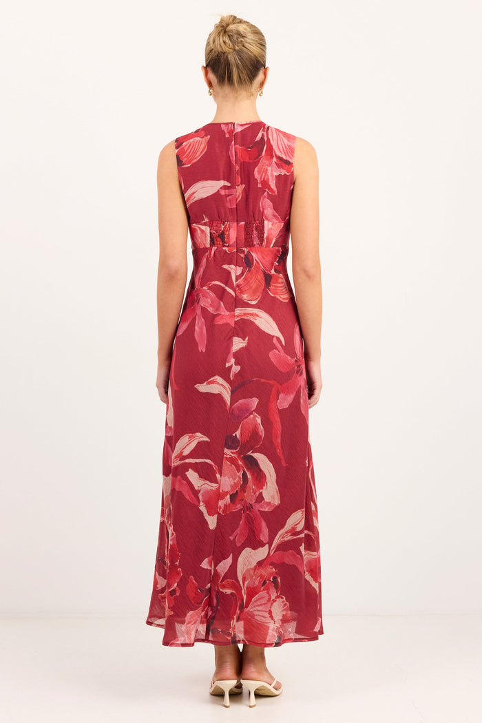 A woman viewed from behind wearing a sleeveless red maxi dress with a large pink and white floral print, standing against a plain white studio background.