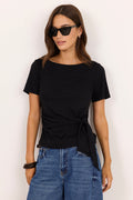 A woman modeling a black short-sleeve boat-neck t-shirt with a twisted front detail, paired with blue jeans, against a plain off-white background.