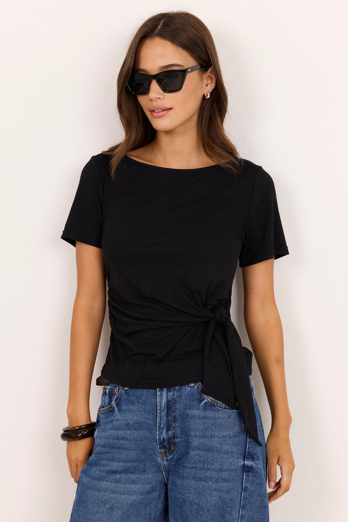 A woman modeling a black short-sleeve boat-neck t-shirt with a twisted front detail, paired with blue jeans, against a plain off-white background.