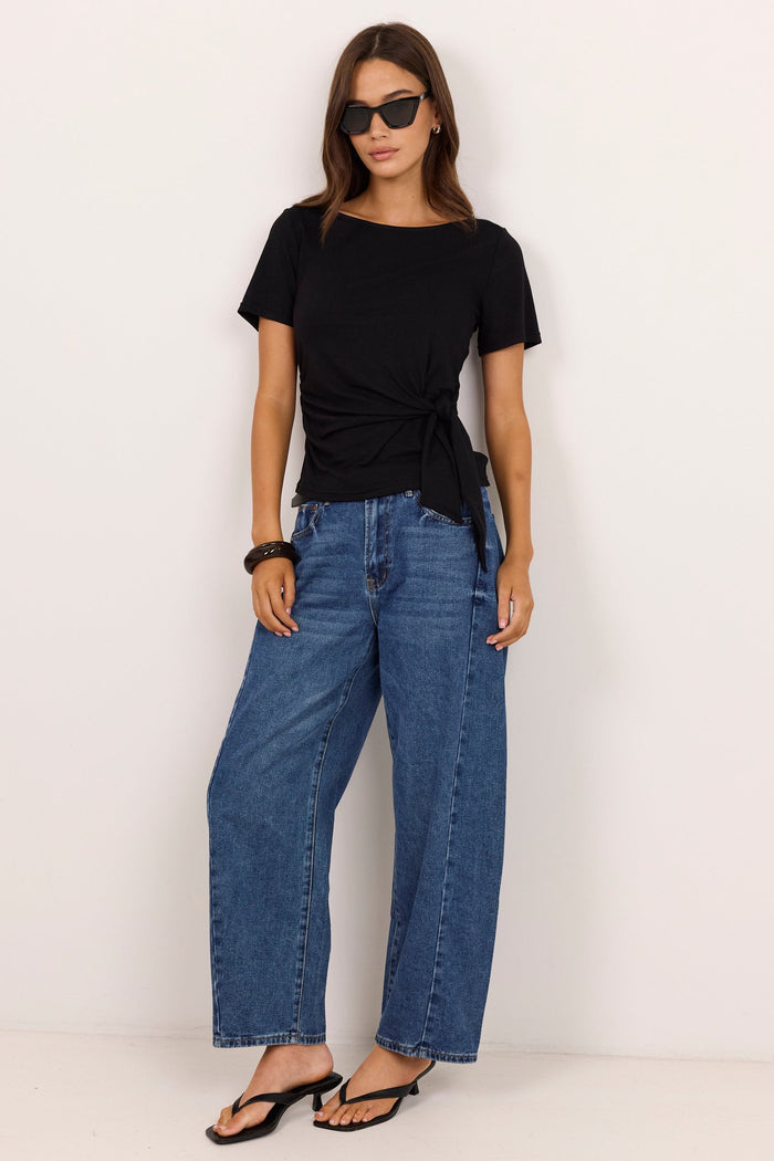 A woman modeling a black short-sleeve t-shirt with a twisted knot detail at the hem, paired with wide-leg blue jeans and black sandals, standing against a plain off-white studio background.