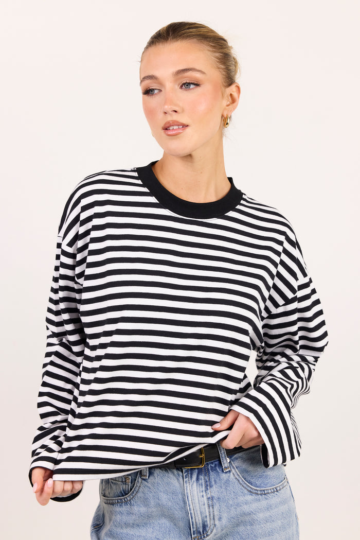A woman wearing a black and white horizontally striped long-sleeve top with a black crew neck, paired with light blue jeans, against a plain light background.