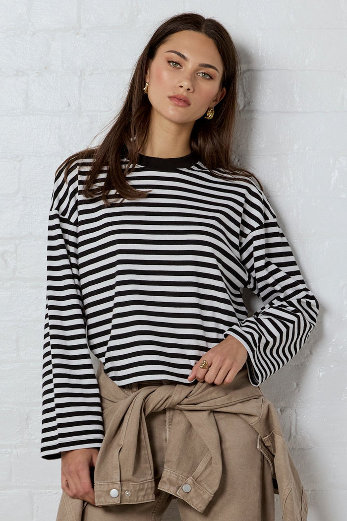 A woman stands against a white brick wall wearing a black and white striped long-sleeve top, beige cropped pants with a matching tie belt, and black flat shoes.