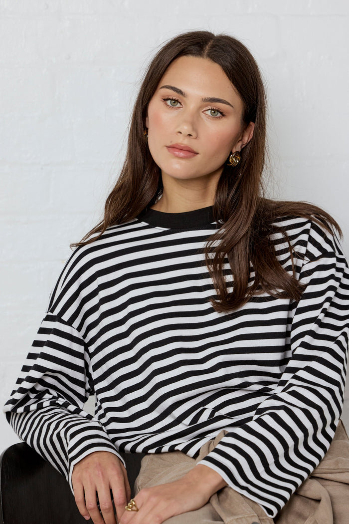 A woman wearing a black and white horizontally striped long-sleeve top, paired with beige pants, sitting against a white brick wall.