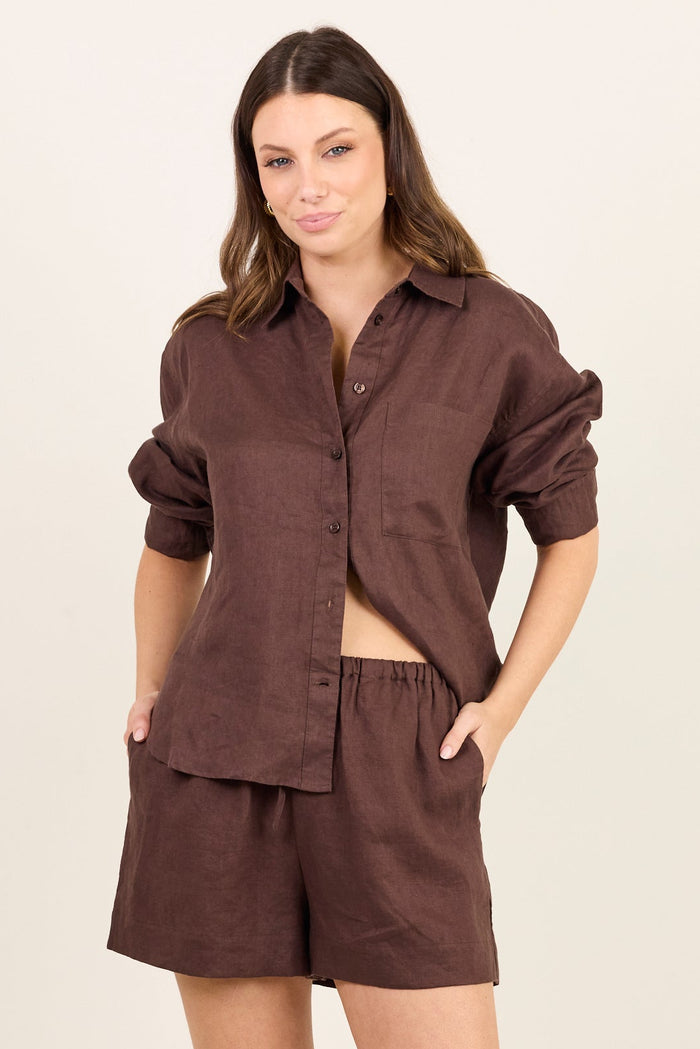 A woman modeling a matching dark brown lounge set consisting of a button-down shirt and shorts, paired with beige flip-flops, against a plain off-white studio background.