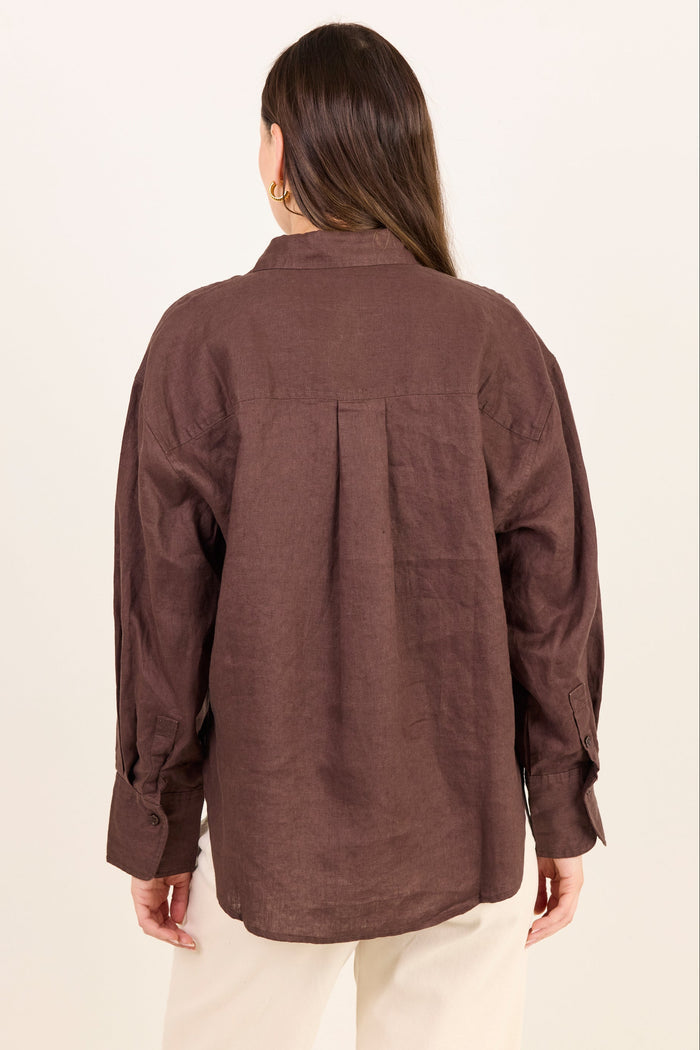 A woman viewed from the back wearing a brown long-sleeve linen shirt and cream pants, standing against a plain off-white background.