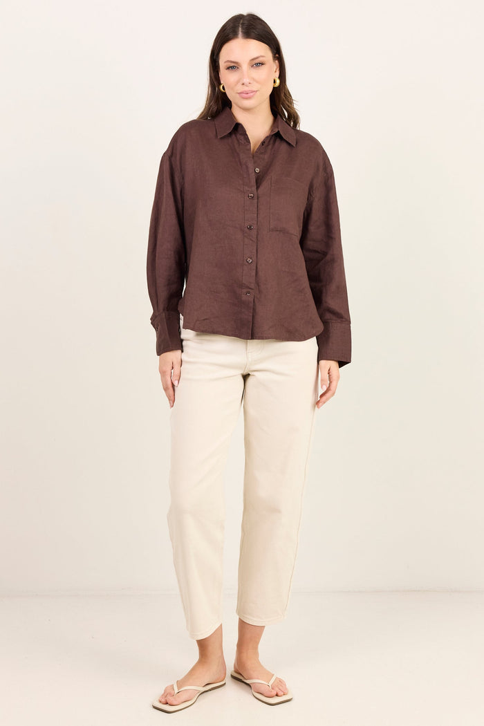 A woman modeling a dark brown long-sleeve button-up shirt and cream cropped trousers, standing against a plain off-white studio background.