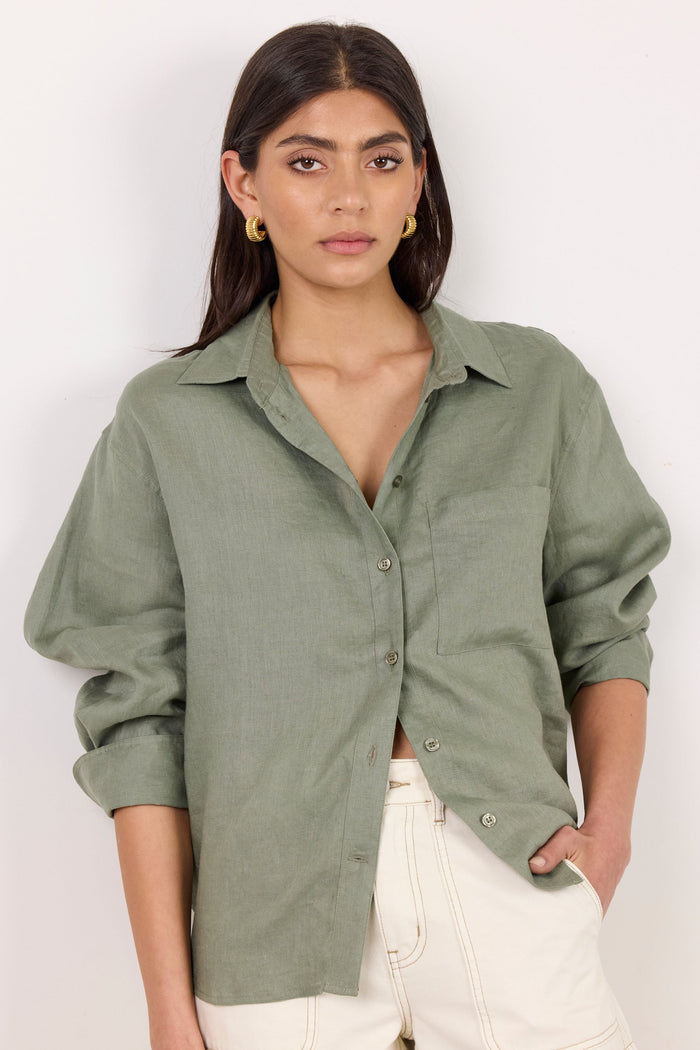 A woman modeling an oversized sage green linen button-up shirt with rolled sleeves and white shorts, set against a plain white studio background.