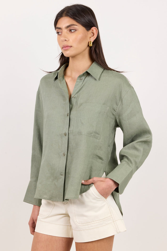 A woman modeling a sage green linen button-up shirt paired with off-white shorts, standing against a plain white studio background.