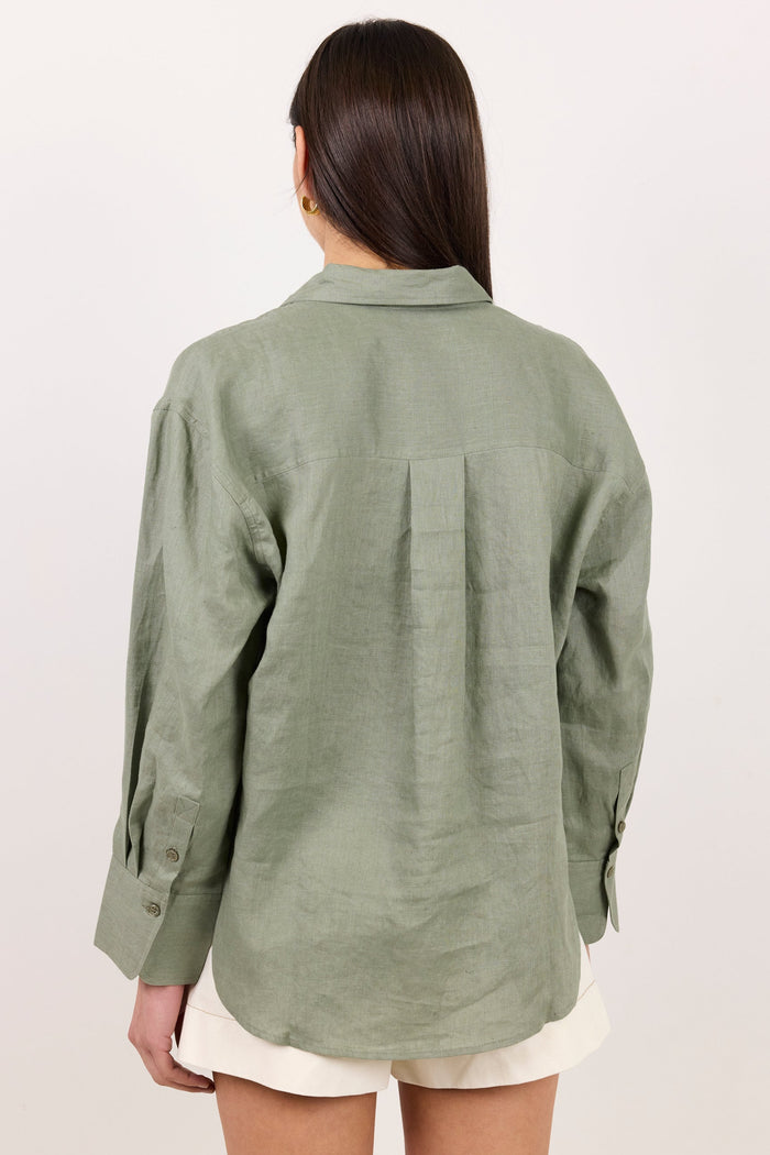 A woman viewed from the back wearing an oversized sage green linen button-up shirt and white shorts against a plain off-white background.