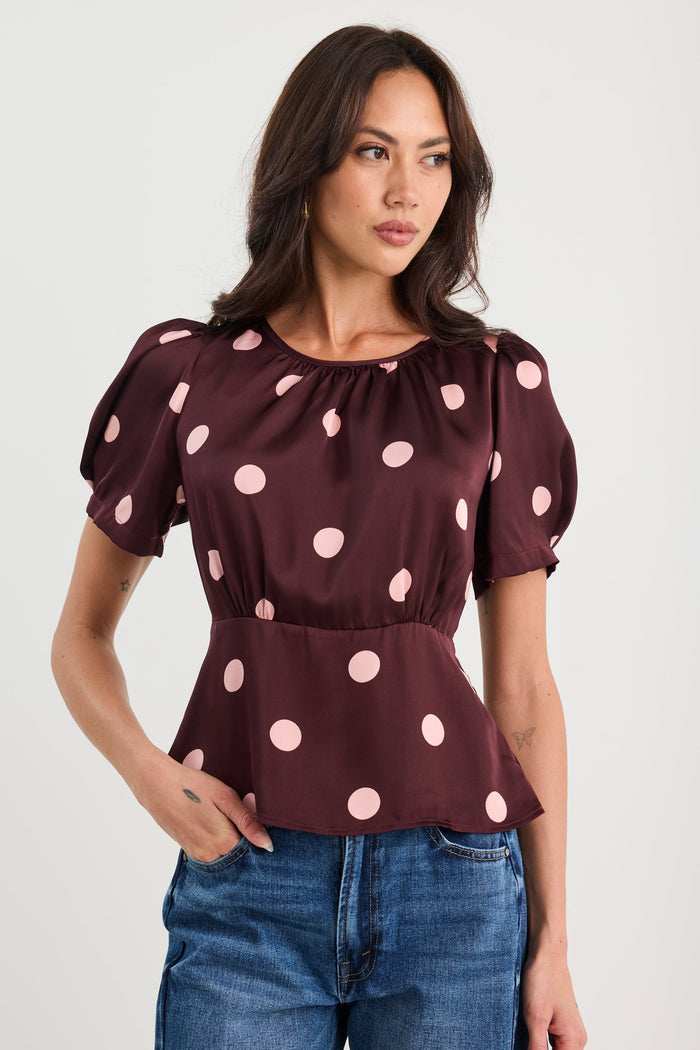 Woman wearing a plum-coloured short sleeve top with blush pink polka dots, gathered neckline, and a cinched waist, styled with blue jeans.
col_pattern