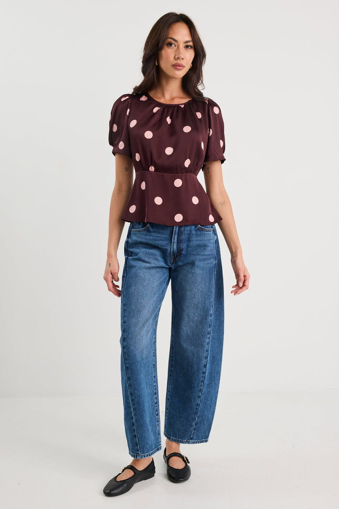 Woman wearing a plum-coloured short sleeve top with blush pink polka dots, gathered neckline, and a cinched waist, styled with blue jeans.