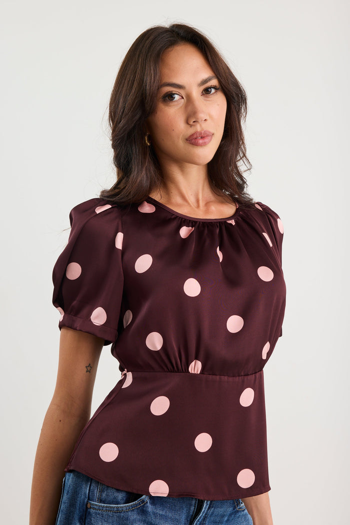 Woman wearing a plum-coloured short sleeve top with blush pink polka dots, gathered neckline, and a cinched waist, styled with blue jeans.
