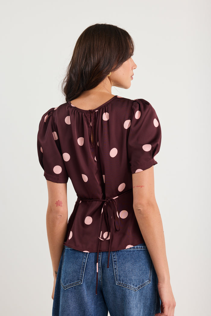 Woman wearing a plum-coloured short sleeve top with blush pink polka dots, gathered neckline, and a cinched waist, styled with blue jeans.