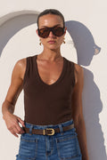 A woman modeling a dark brown ribbed tank top and blue jeans with a brown belt, standing against a white stucco wall with an arched recess.