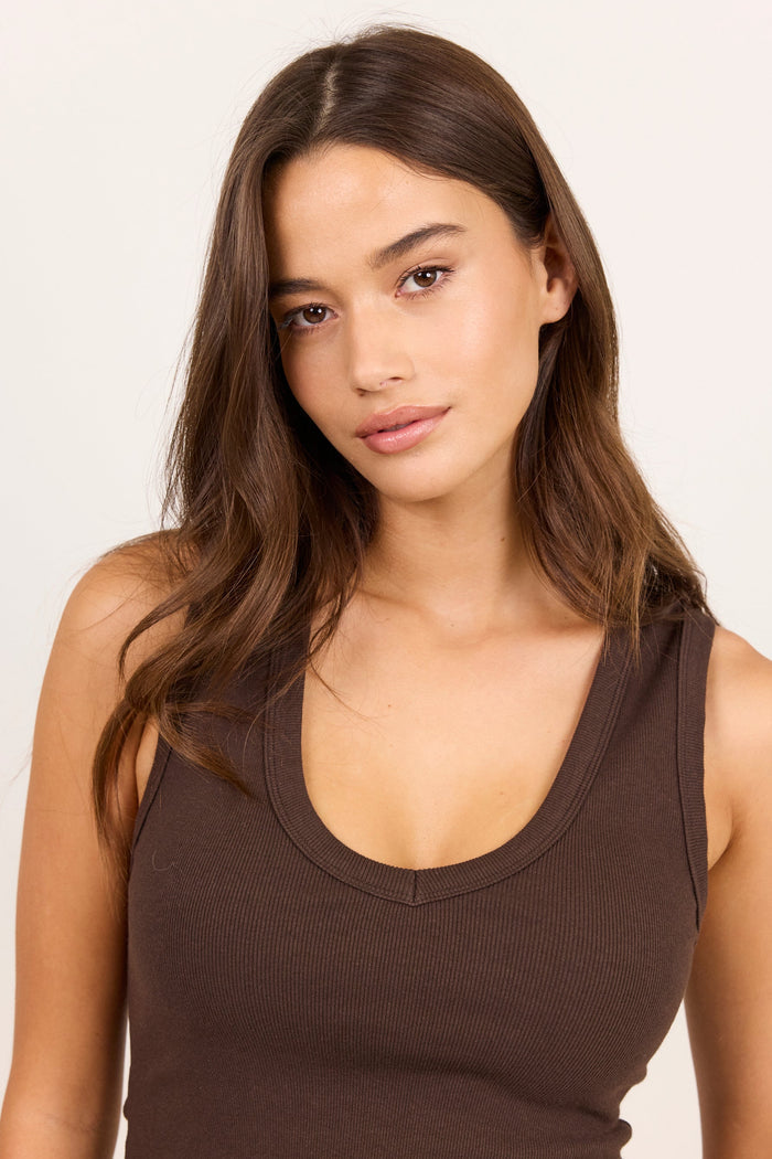 A woman with long brown hair wearing a dark brown ribbed scoop-neck tank top, photographed against a clean off-white studio background.