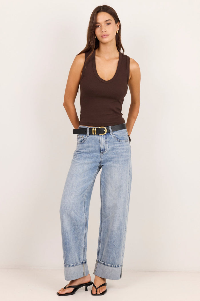 A woman modeling a dark brown ribbed tank top, wide-leg light-wash blue jeans with rolled hems, a black belt with a gold buckle, and black heeled sandals, standing against a plain off-white studio background.