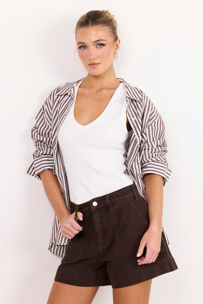 A woman modeling a casual outfit consisting of a white V-neck tank top, dark brown shorts, and a brown and white striped button-up shirt draped off her shoulders, set against a plain white background.
