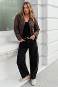 Woman standing against a white building wearing a brown jacket and black pants.