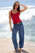 A woman stands on a concrete pier by the ocean wearing a bright red tank top, wide-leg blue jeans, and black sandals, with waves and blue sky in the background.