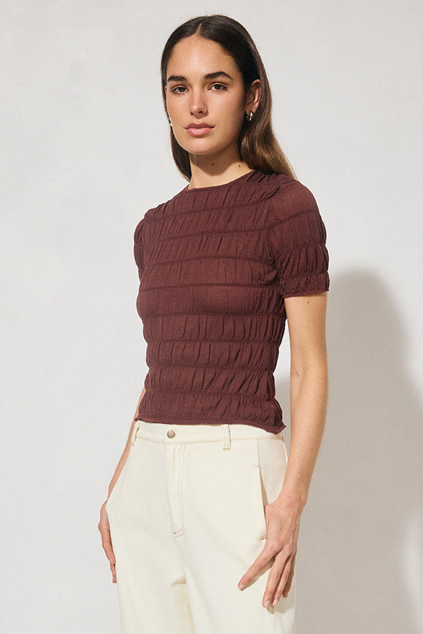 Woman wearing a brown ribbed sweater and white pants against a plain background